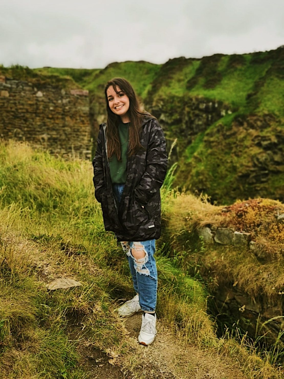 A young woman is smiling outdoors, standing on grassy terrain with rocky ruins and green hills in the background. She wears a dark jacket, green top, ripped jeans, and white sneakers. The sky is overcast.