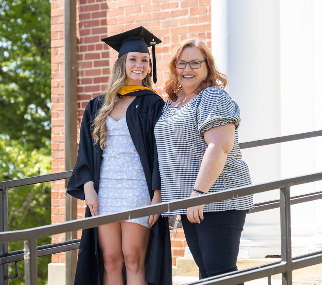 A young woman in a graduation cap and gown stands smiling next to an older woman in glasses and a checkered blouse, posing outdoors by a brick building and white column.