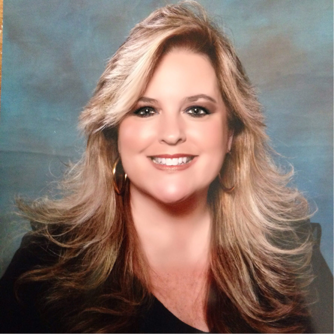 Smiling woman with long, voluminous blonde hair, wearing hoop earrings and a black top, posing in front of a blue and gray studio background.