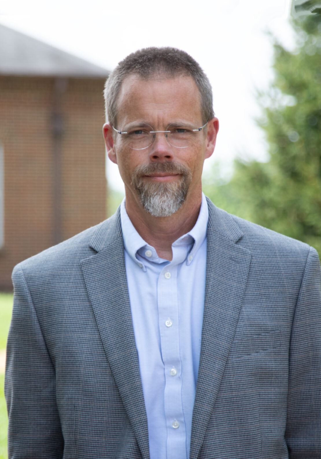 A middle-aged man with glasses, short hair, and a beard, wearing a light blue dress shirt and a gray blazer, stands outdoors with a blurred building and greenery in the background.