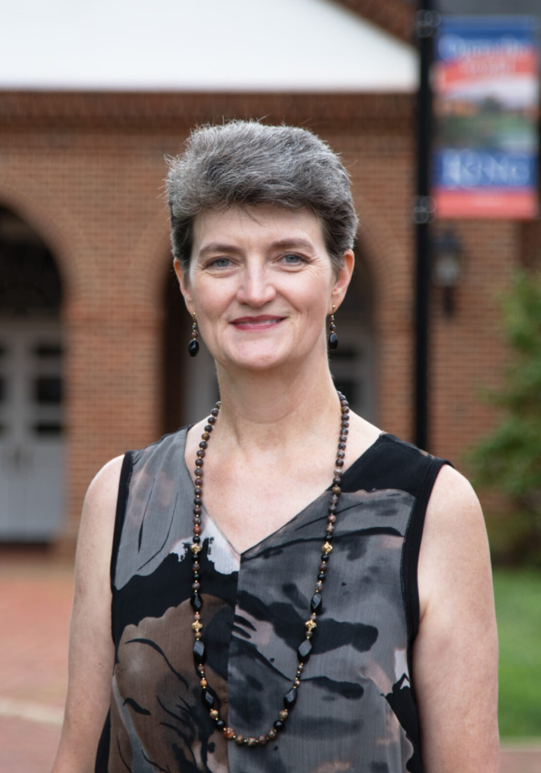 A woman with short gray hair, wearing a sleeveless patterned top and a beaded necklace, stands outdoors in front of a brick building with arched doorways and a blurred sign in the background.