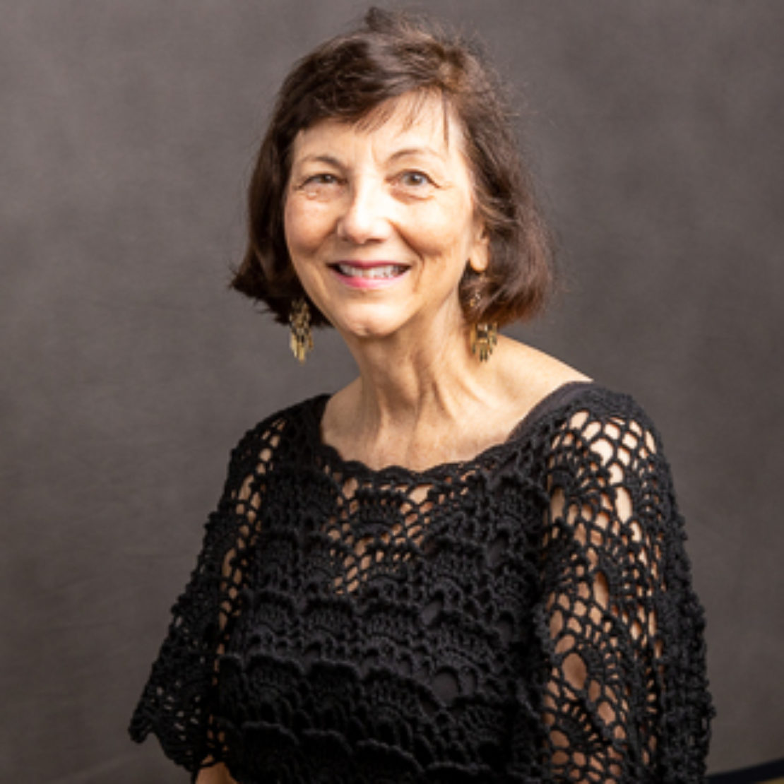 An older woman with short dark hair smiles at the camera. She wears gold earrings and a black, lacy, crocheted top. The background is plain and dark.