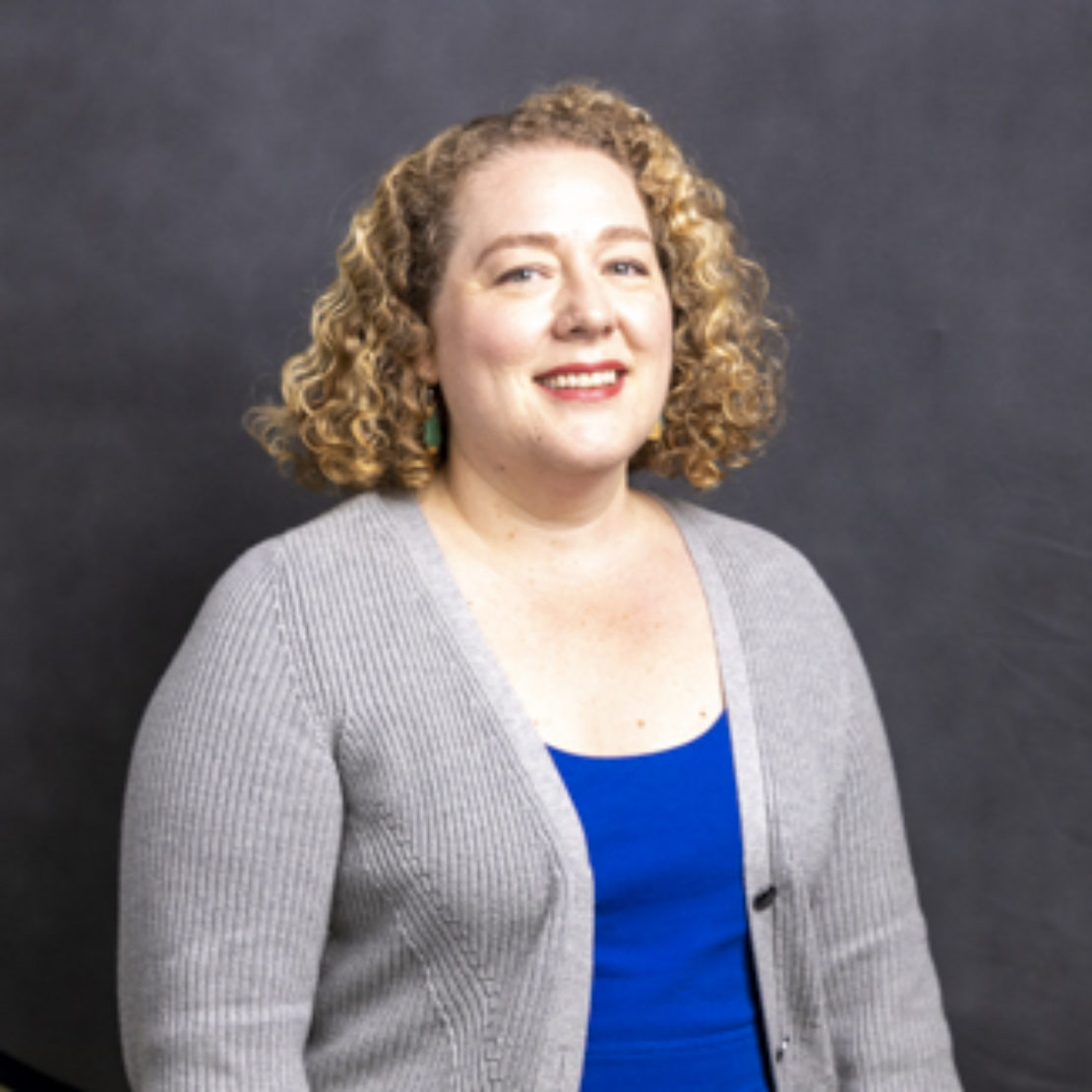 A woman with curly blonde hair, wearing a gray cardigan over a blue top, stands in front of a plain dark background, smiling at the camera.