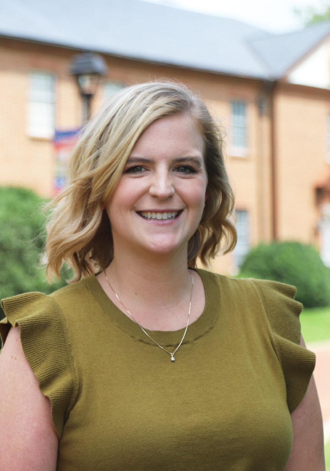 A woman with shoulder-length blonde hair, wearing an olive green top and a necklace, smiles while standing outdoors in front of a brick building and greenery.