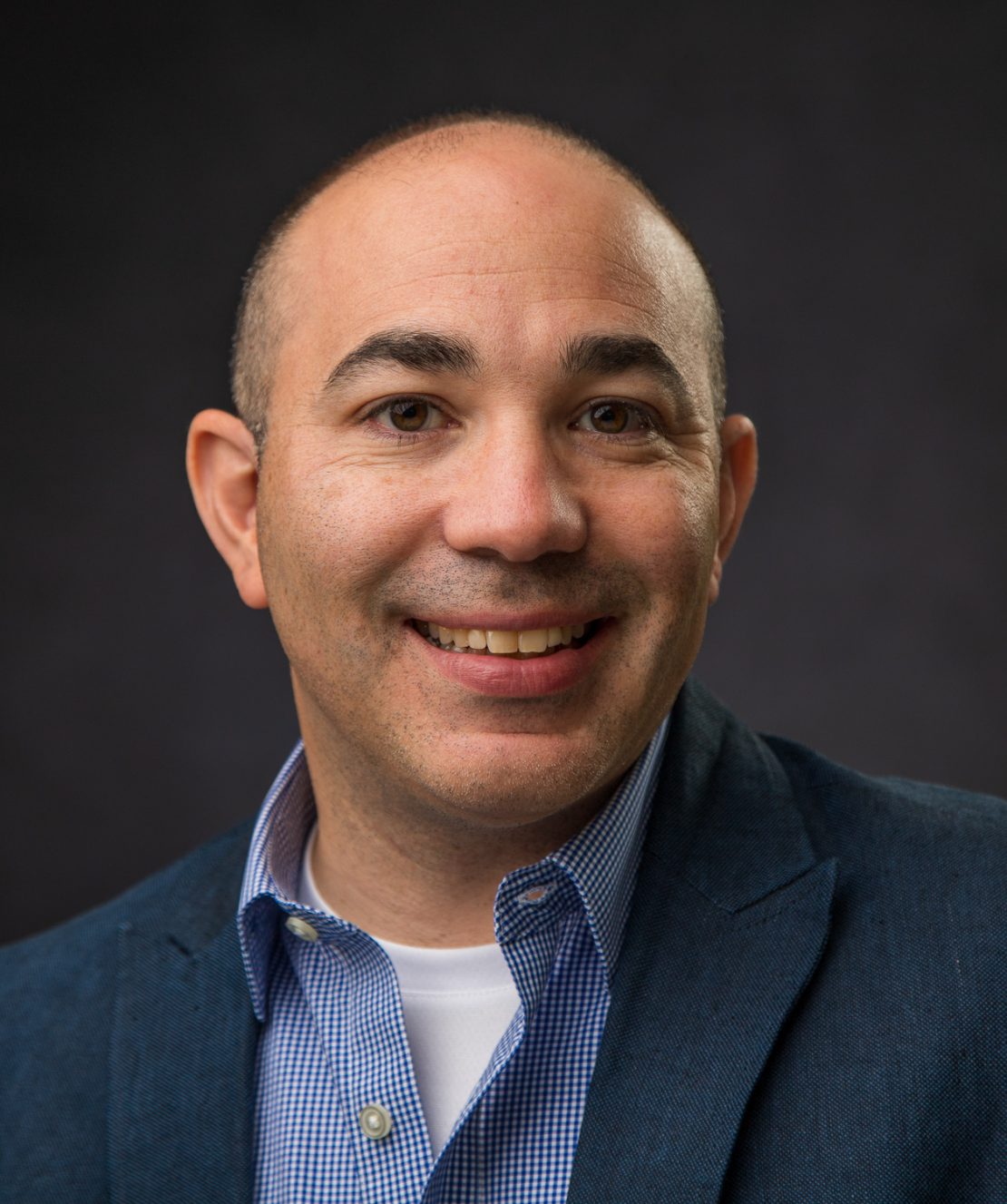 A smiling man with short hair, wearing a blue blazer over a light blue checked shirt and white undershirt, poses in front of a dark, blurred background.