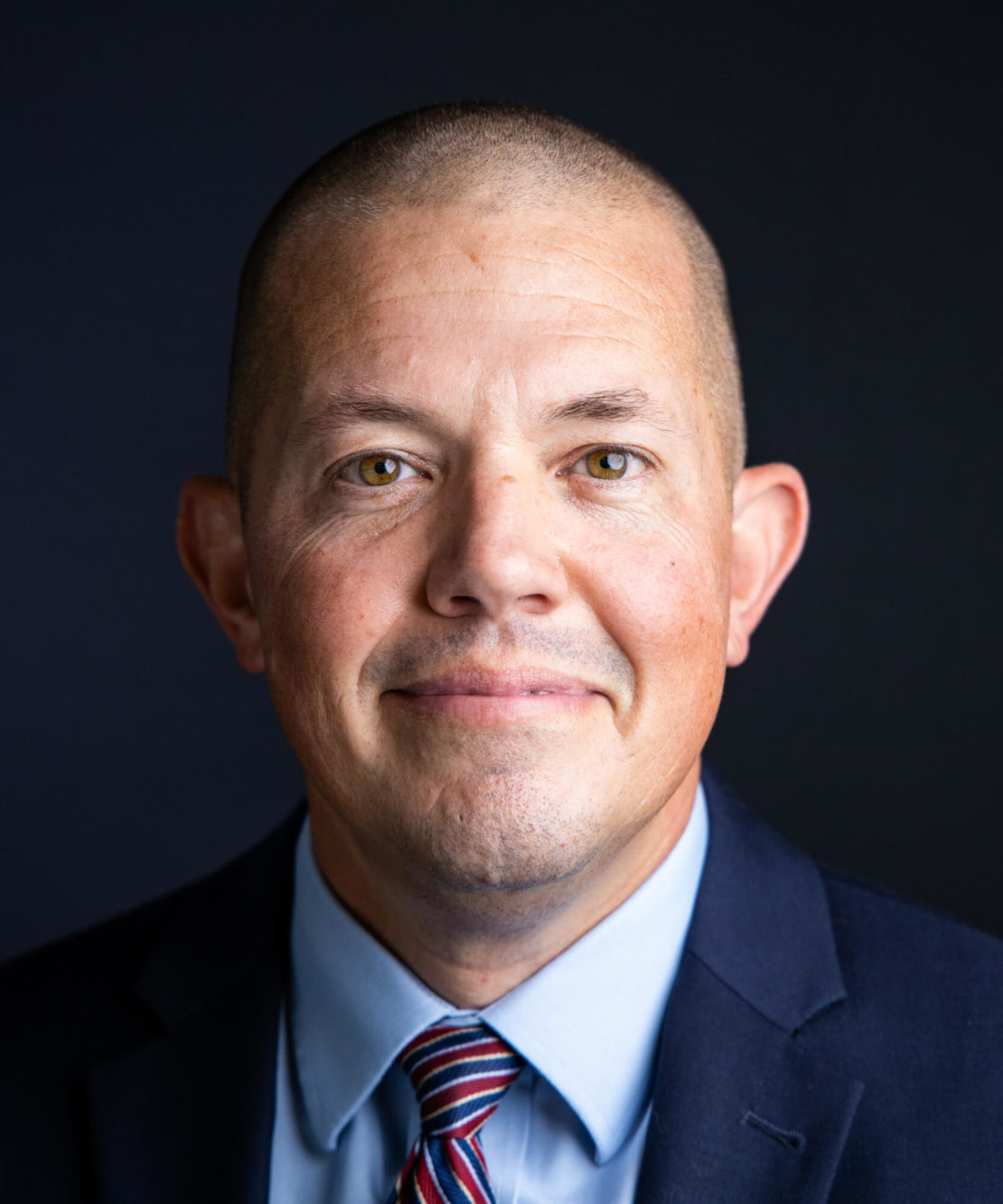 A man with closely shaved hair wearing a dark suit, light blue shirt, and striped tie, smiling slightly in front of a dark background.