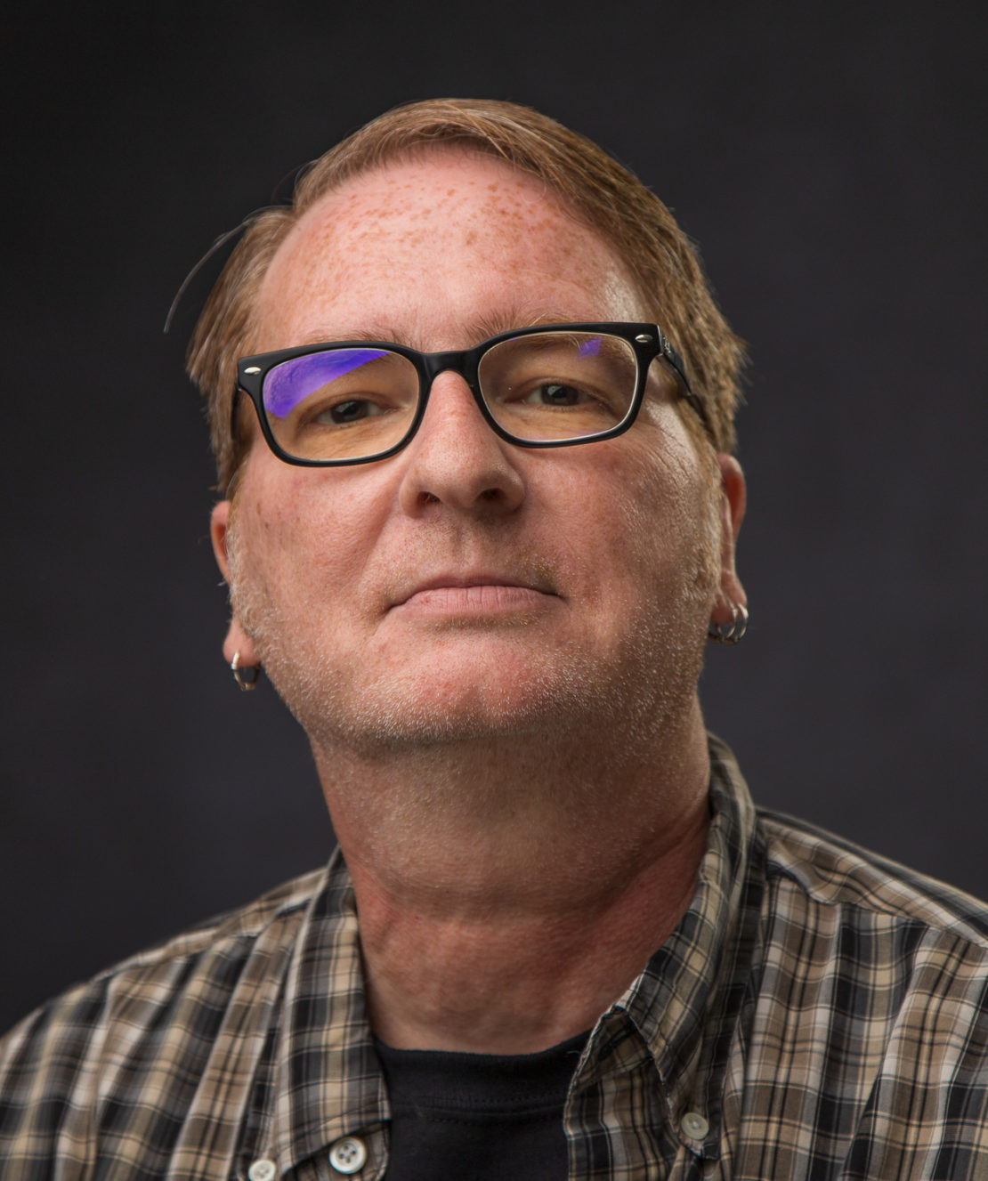 A person with short light brown hair, wearing black-rimmed glasses, earrings, and a plaid shirt, looks confidently at the camera against a dark background.