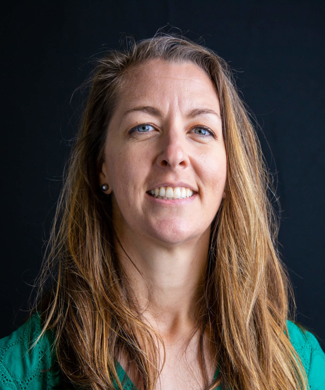 A woman with long brown hair and blue eyes smiles softly at the camera. She is wearing a green top and small round earrings, with a dark, solid background behind her.