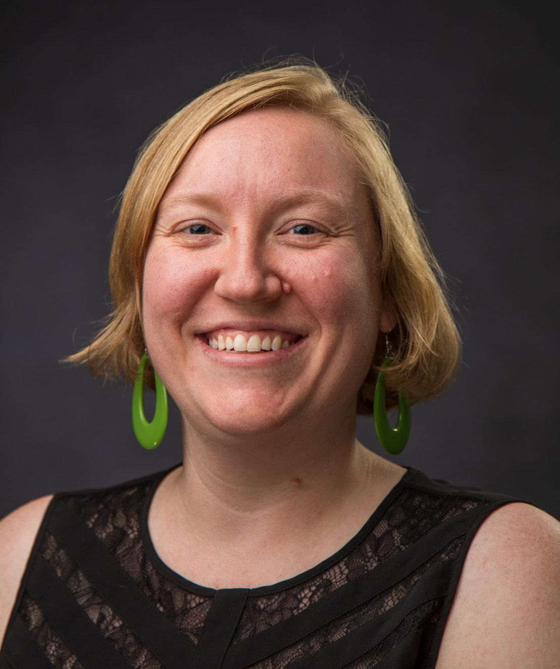 A woman with short blonde hair smiles at the camera. She is wearing large green hoop earrings and a sleeveless black top with a lace panel. The background is dark and blurred.