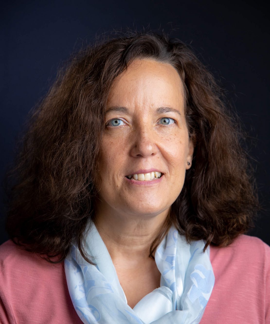 A woman with curly brown hair, blue eyes, and light skin smiles at the camera. She is wearing a pink top and a light blue patterned scarf, posed against a dark background.