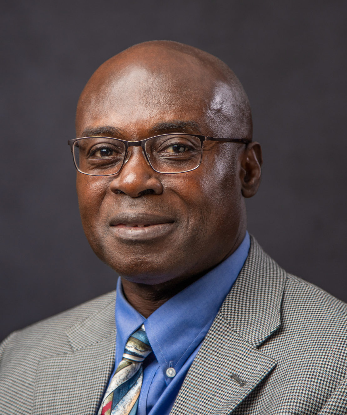 A man wearing glasses, a gray checked suit jacket, a blue shirt, and a multicolored tie poses against a dark, plain background, looking calmly at the camera.