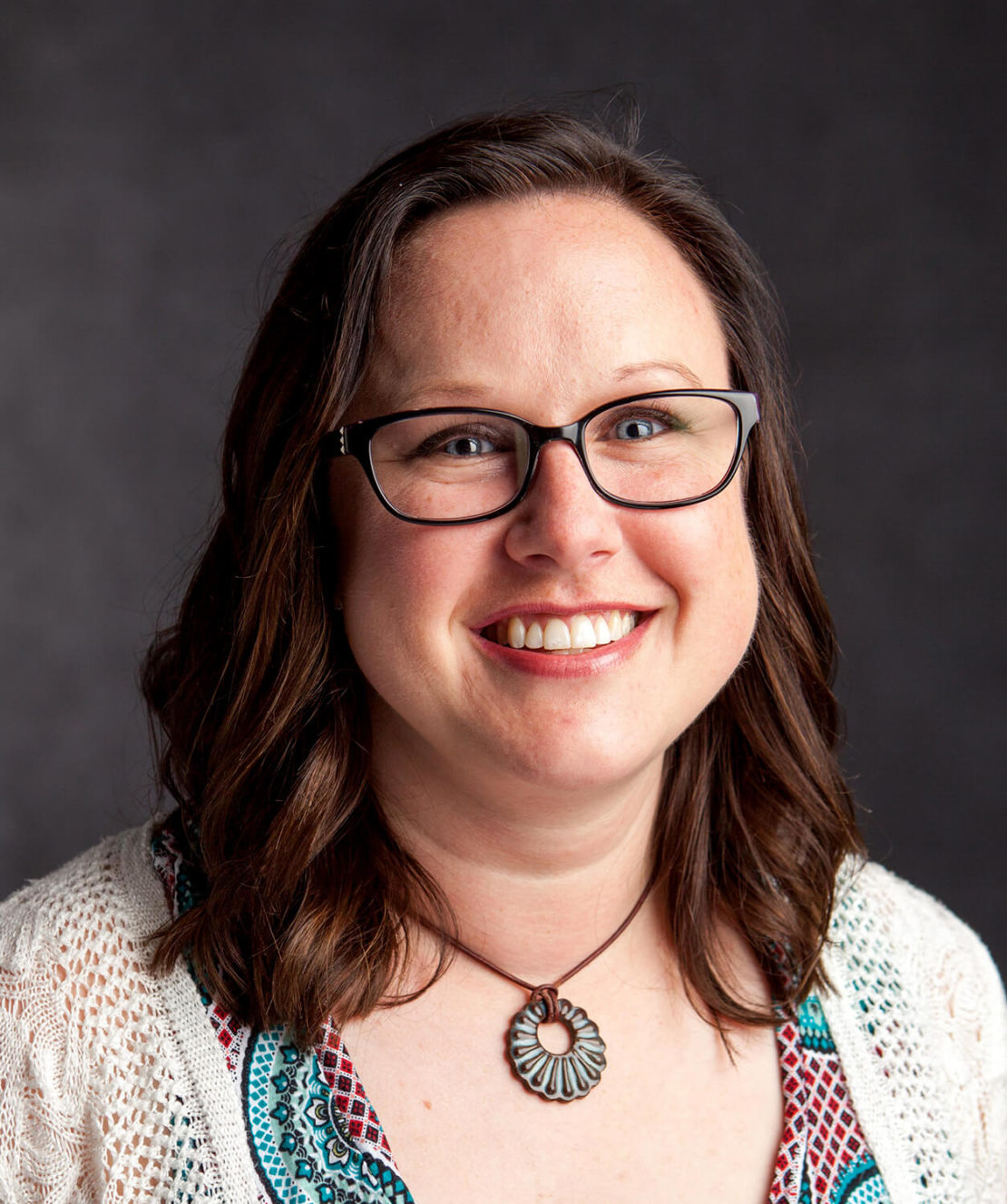 A smiling woman with shoulder-length brown hair, wearing glasses, a white patterned top, and a circular pendant necklace, poses in front of a dark background.