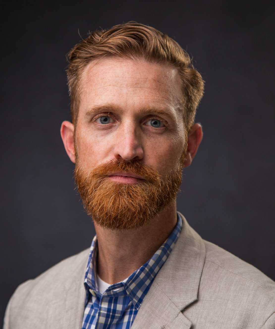 A man with red hair and a beard wearing a light beige blazer over a blue plaid shirt poses against a dark, plain background.