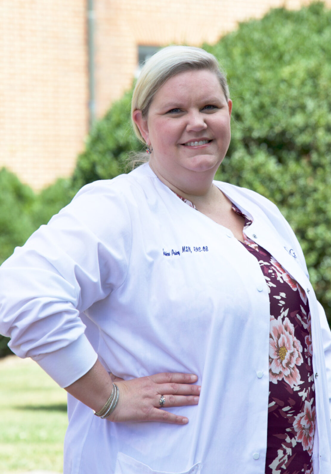A woman with blond hair pulled back, wearing a white medical coat over a floral top, stands outside in front of greenery and a brick building, smiling at the camera with her hand on her hip.