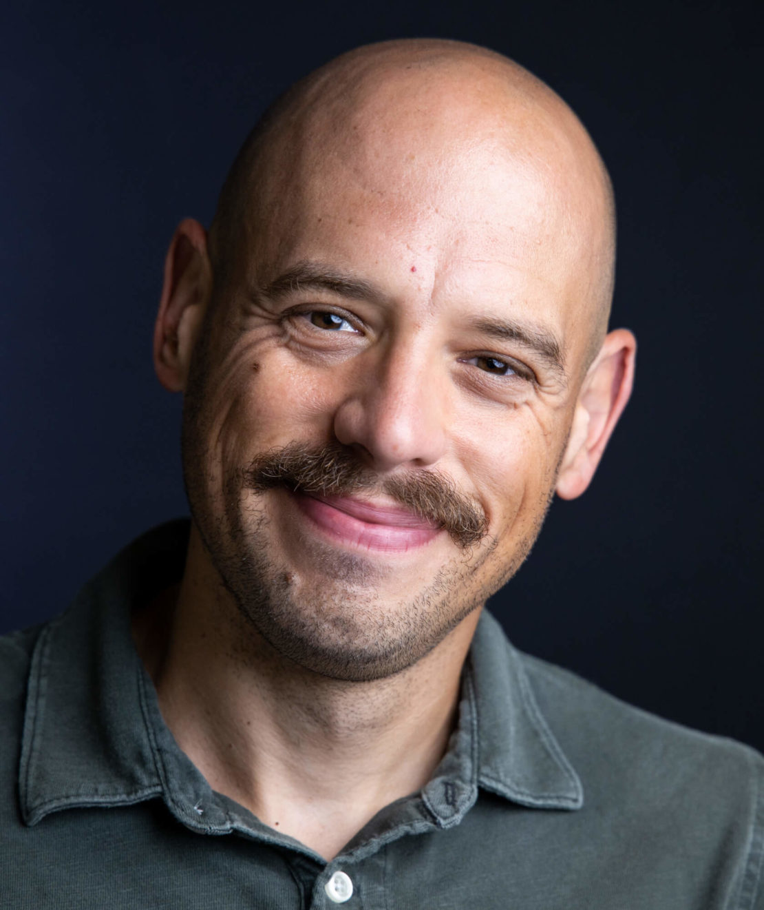 A smiling bald man with a mustache wearing a green collared shirt poses against a dark background.