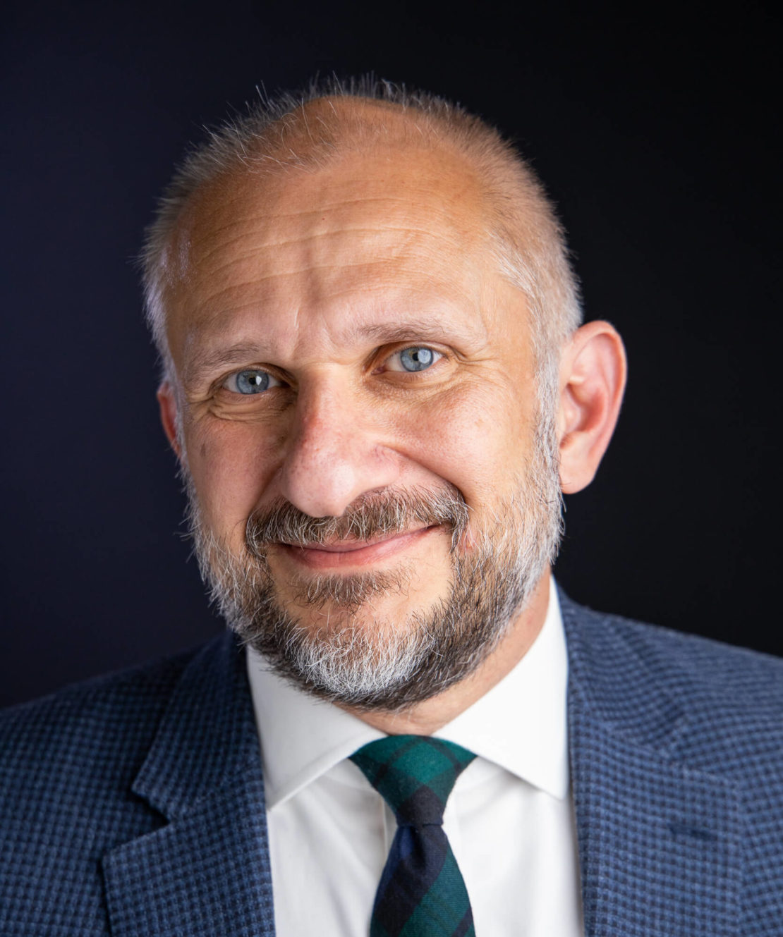 A middle-aged man with a light beard and short hair smiles, wearing a blue checkered suit jacket, white shirt, and striped tie, against a dark background.