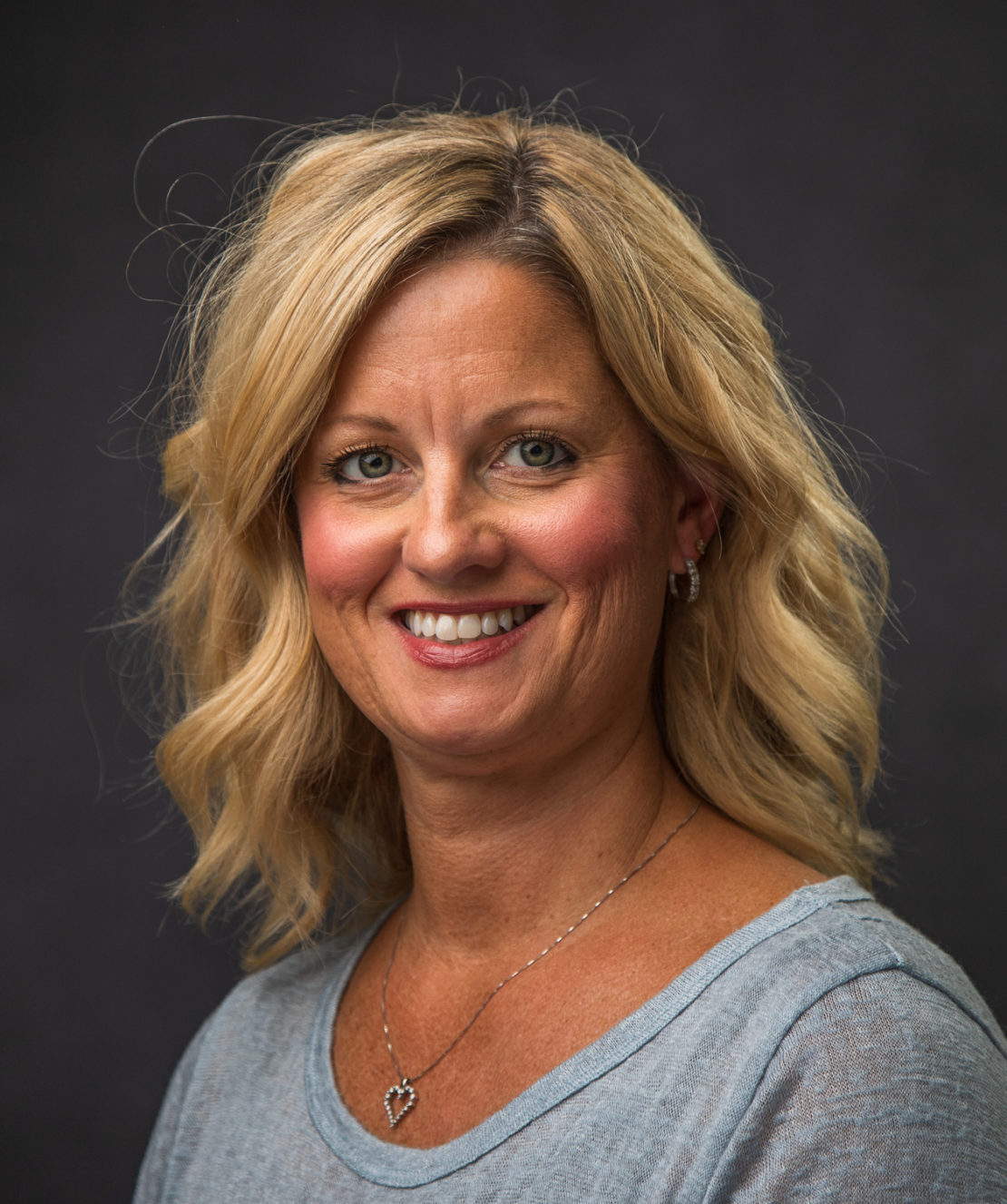 A woman with shoulder-length blonde hair and a light blue top smiles at the camera against a dark gray background. She wears a heart-shaped necklace and small earrings.