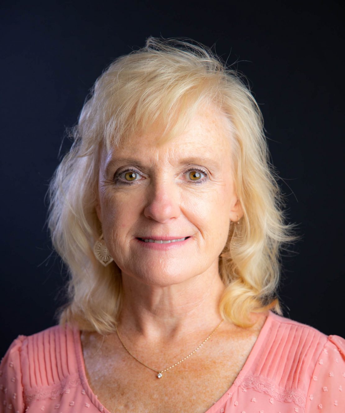 A middle-aged woman with blonde, shoulder-length hair is wearing a light pink top and a delicate necklace. She is smiling slightly and looking directly at the camera. The background is plain and dark.