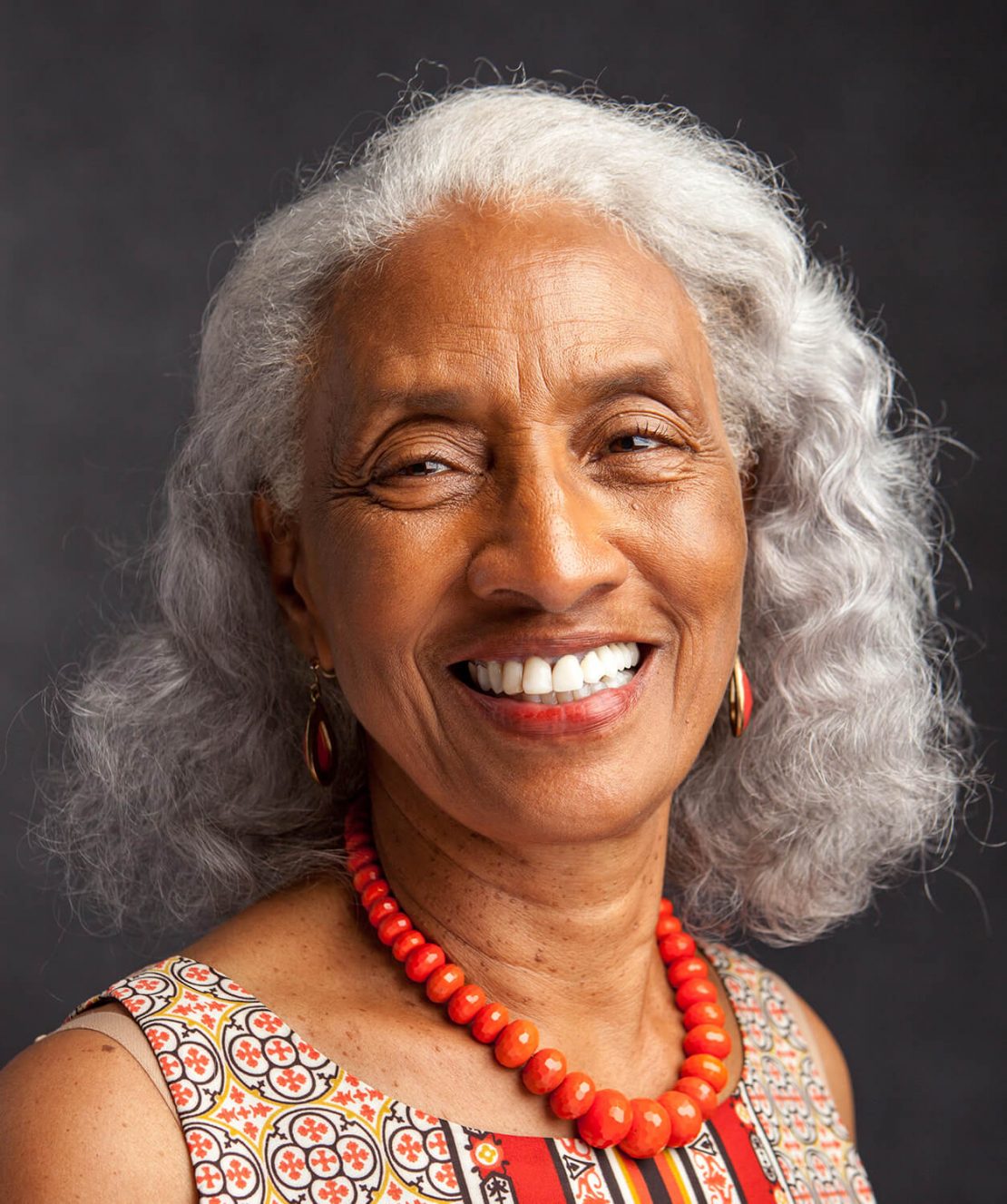 Smiling older woman with gray curly hair, wearing a colorful patterned top, red beaded necklace, and matching earrings, against a dark background.