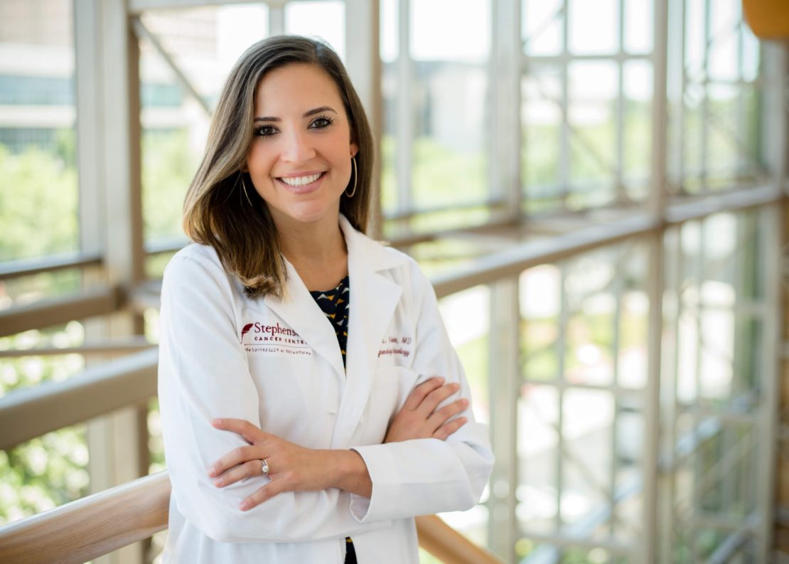A smiling woman with shoulder-length brown hair stands with arms crossed, wearing a white lab coat, in a bright, modern building with large windows and natural light.