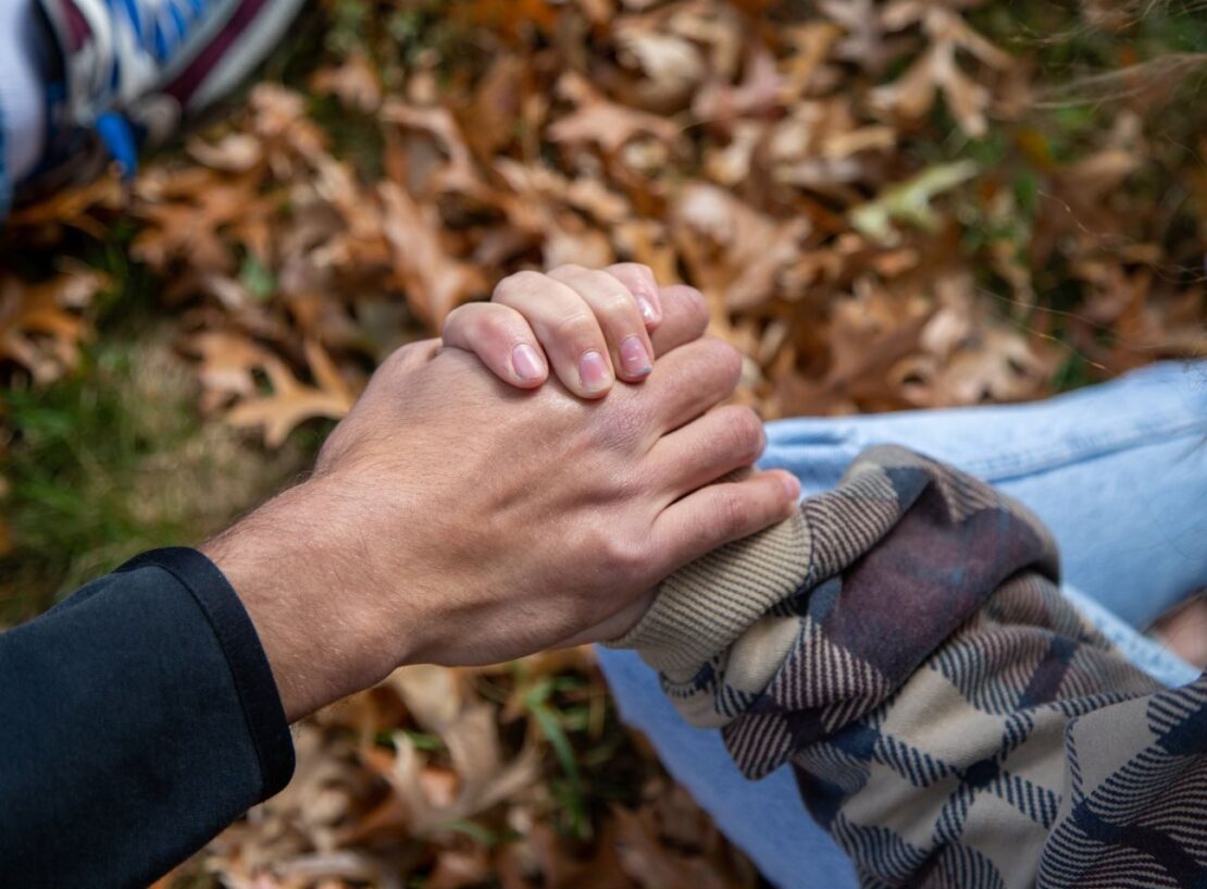 Two people are holding hands outdoors above fallen autumn leaves. One person is wearing a plaid jacket, the other a black sleeve. Part of a sneaker and blue jeans are visible in the background.