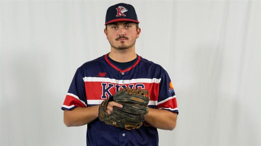 A baseball player in a blue and red uniform and cap stands against a plain white background, holding a brown glove in front of his chest and looking at the camera.