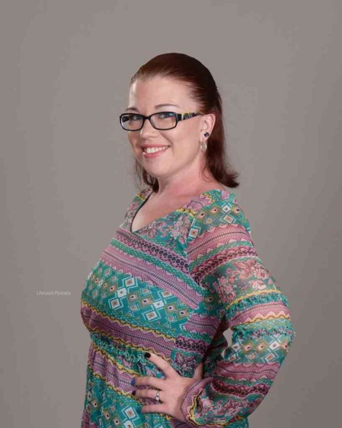 A woman with glasses and reddish-brown hair pulled back smiles while posing with her hand on her hip, wearing a colorful patterned dress against a plain gray background.