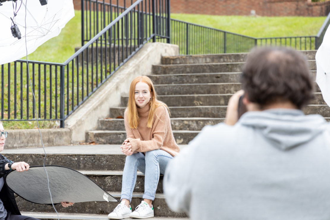 A young woman with long red hair sits on outdoor steps, smiling at the camera. She wears a tan sweater and light jeans. A photographer and an assistant with a reflector are visible in the foreground.