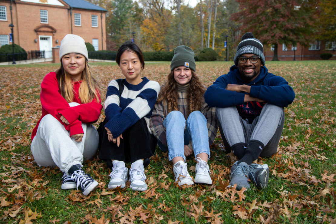 Four college students sit cross-legged on grass covered with autumn leaves, smiling at the camera. Brick buildings and trees with fall foliage are in the background.