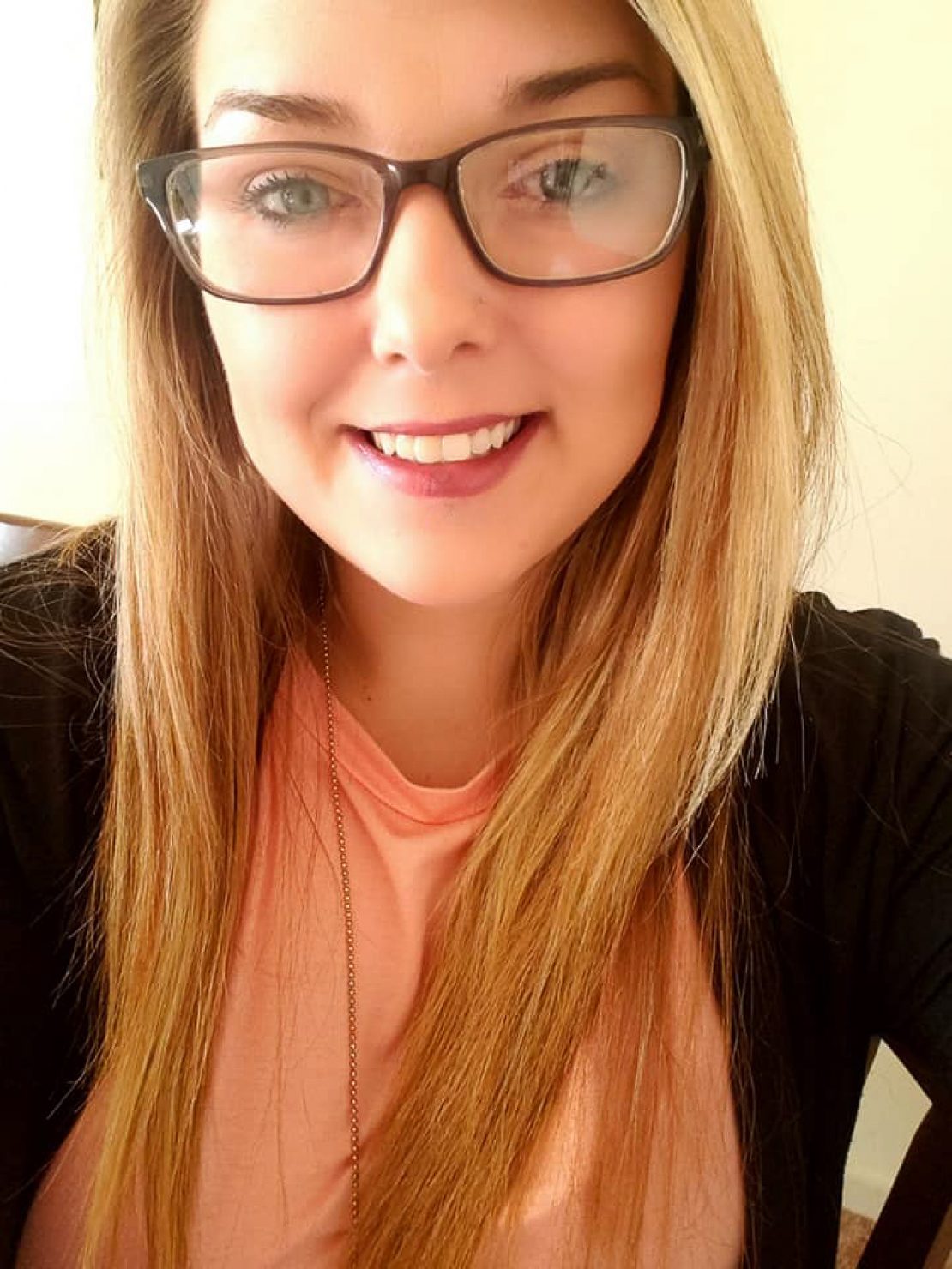 A young woman with long blonde hair and glasses smiles at the camera. She is wearing a light pink top and a black sweater, with a gold necklace. The background is softly lit.