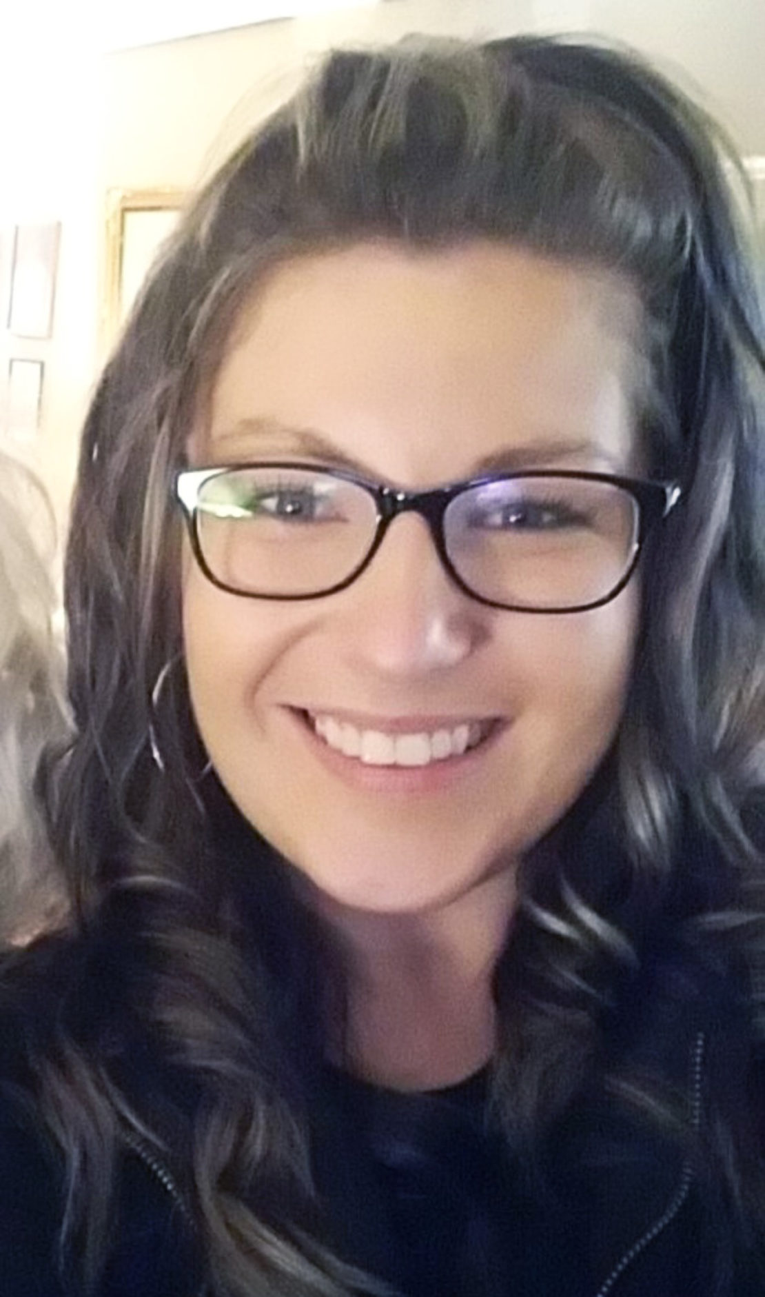 A woman with long, wavy brown hair and glasses smiles at the camera. She is wearing hoop earrings and a dark top, and appears to be indoors.