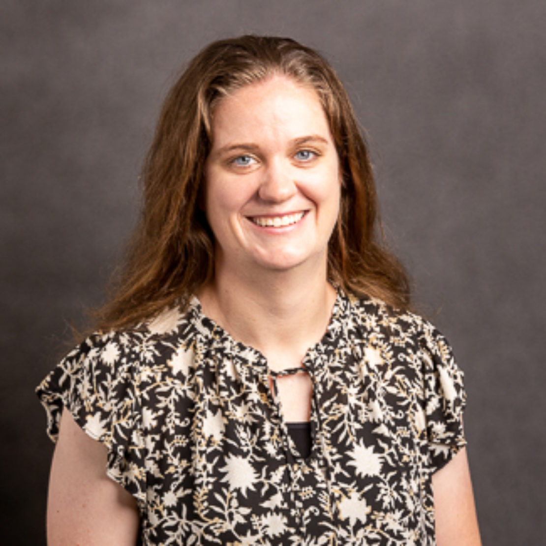 A woman with long brown hair smiles at the camera. She is wearing a black and white floral-patterned blouse and is standing in front of a plain, dark gray background.
