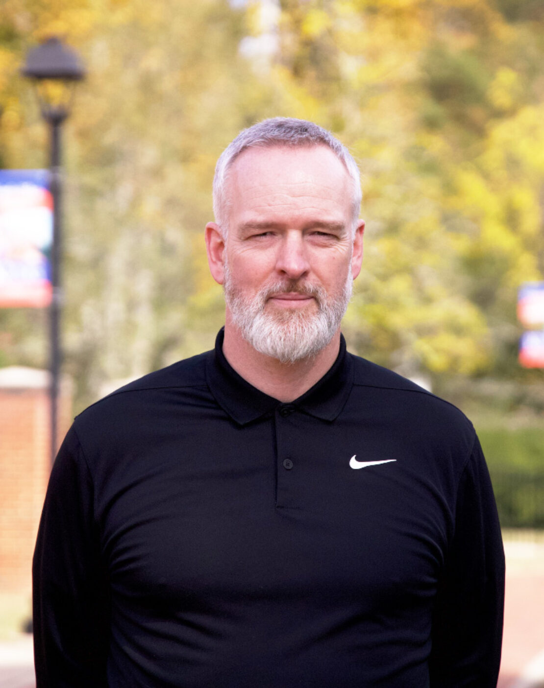 A man with short gray hair and a beard stands outdoors, wearing a black Nike collared shirt. The background is blurred with greenery and some signs visible.