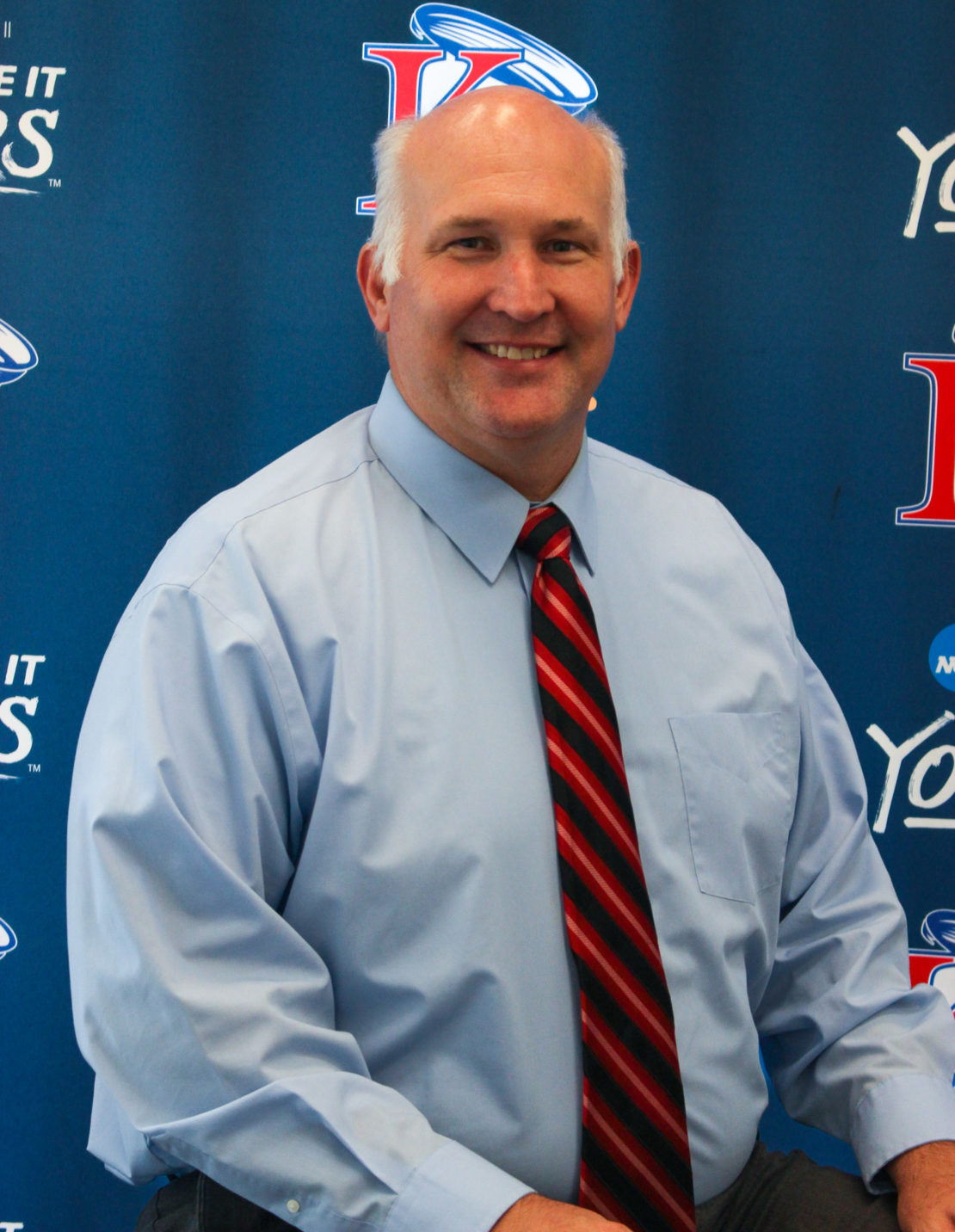 A man with short gray hair, wearing a light blue dress shirt and a red striped tie, smiles while seated in front of a blue backdrop with sports team logos.