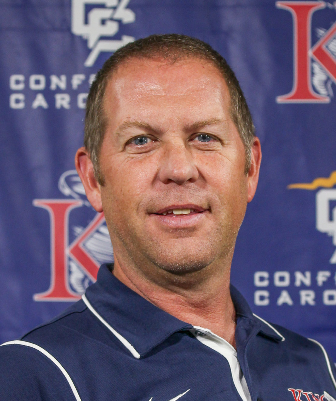 A man with short light brown hair, wearing a navy blue collared shirt with white piping, stands in front of a blue backdrop with red, white, and yellow logos and text.