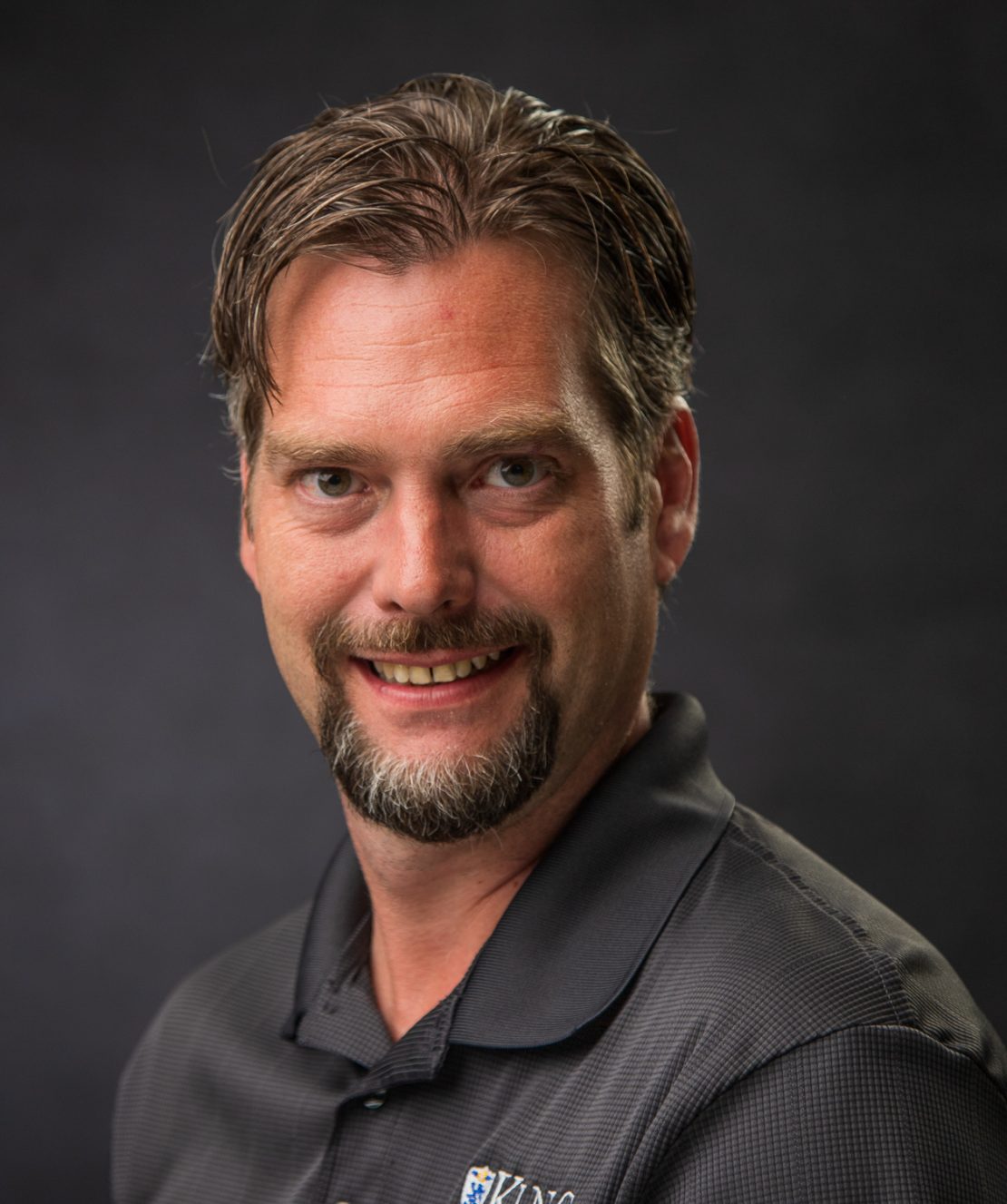 A man with short brown hair, a goatee, and mustache smiles at the camera. He wears a dark collared shirt with a small logo on it, and the background is dark and plain.