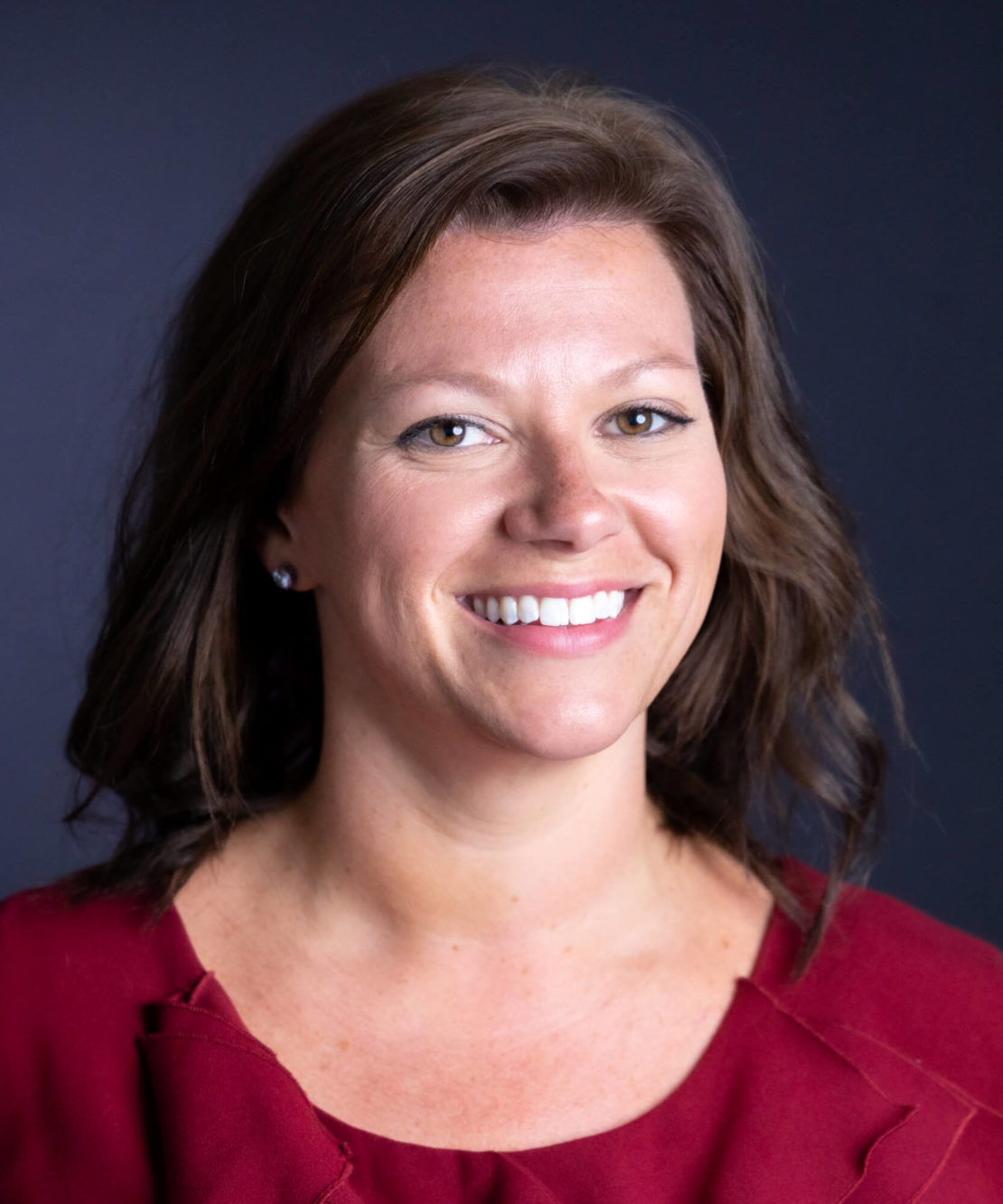 A woman with medium-length brown hair smiles at the camera. She is wearing a red top and small earrings, with a dark, plain background behind her.