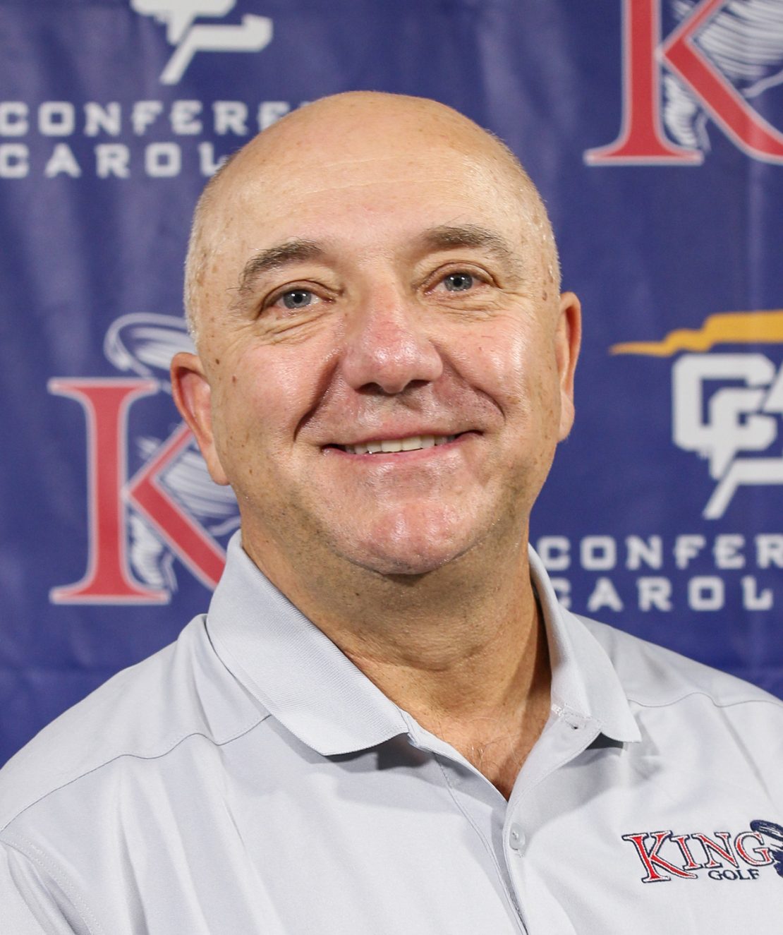 A smiling bald man wearing a light gray shirt with a King Golf logo stands in front of a blue backdrop displaying Conference Carolinas and various sports logos.