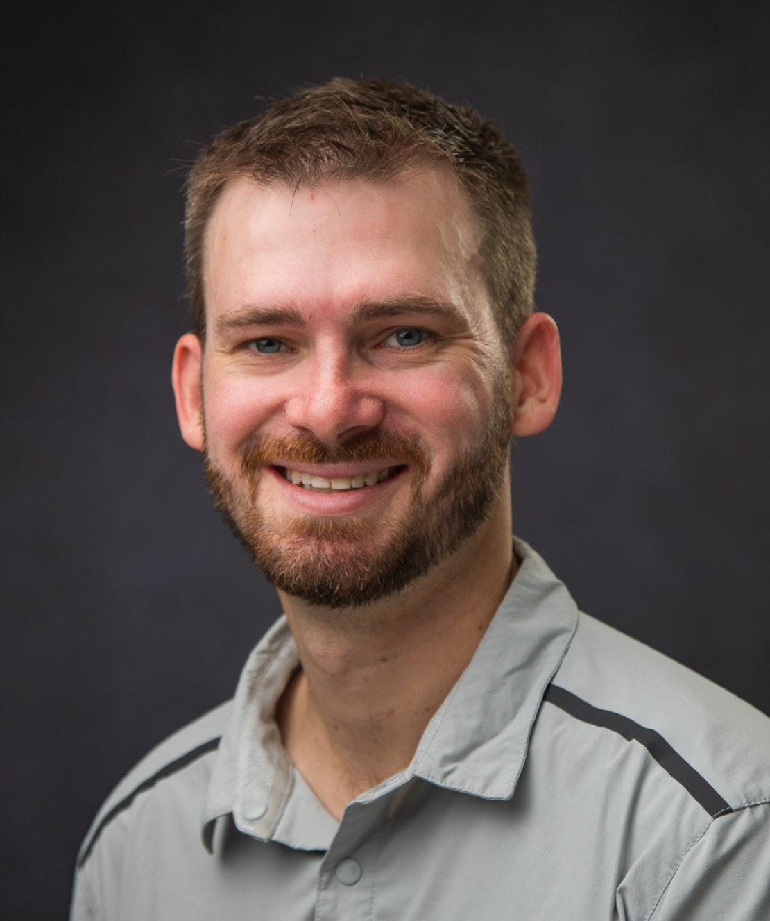 A man with short brown hair and a beard smiles at the camera. He is wearing a light gray collared shirt and is posed against a dark, plain background.