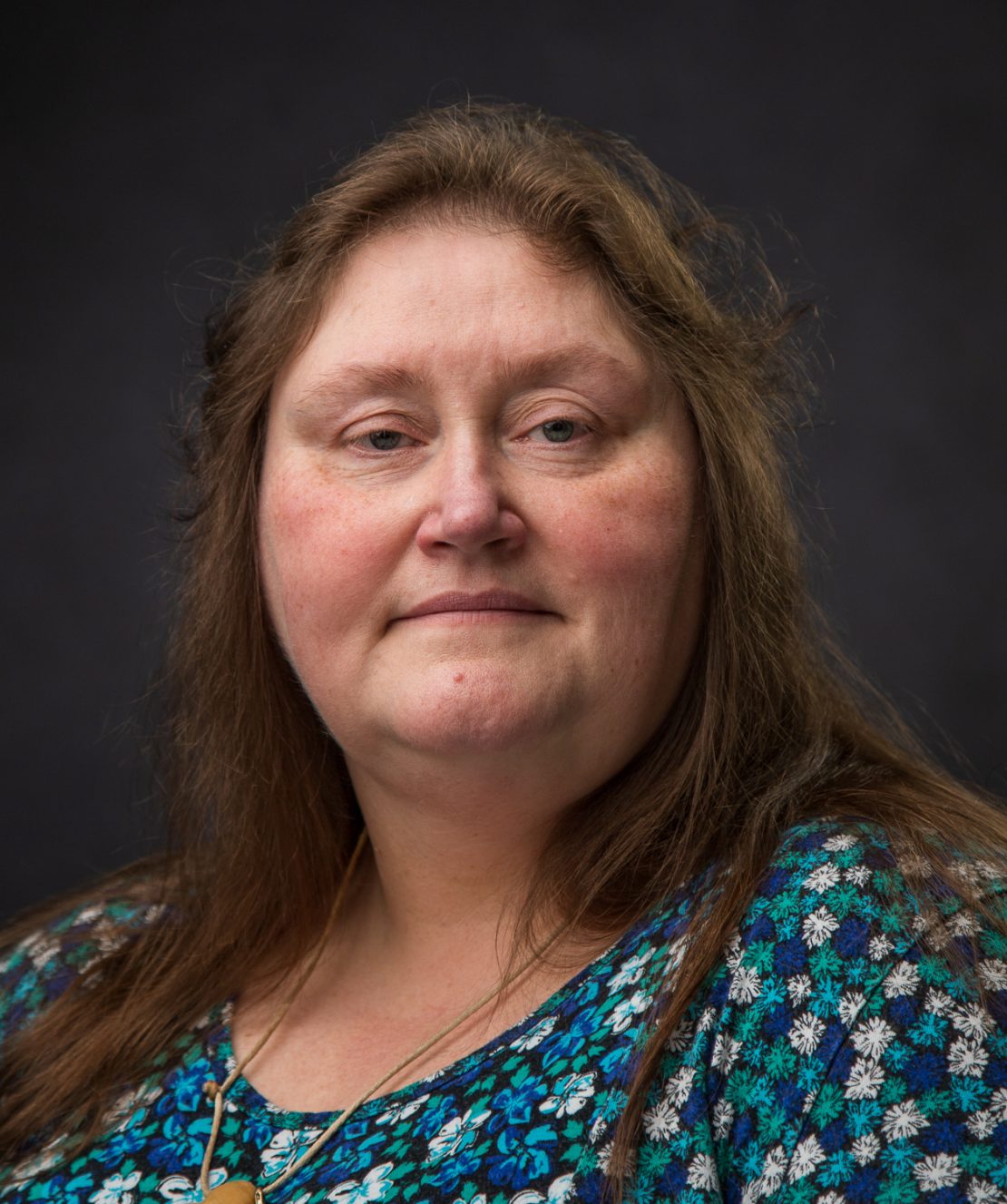 A woman with long brown hair wearing a blue patterned top poses in front of a dark, plain background, looking directly at the camera with a neutral expression.