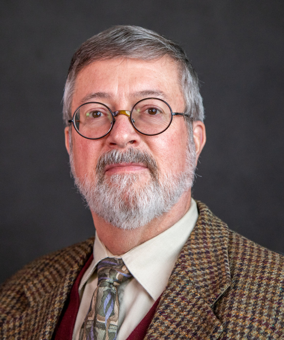 A middle-aged man with gray hair and beard, wearing round glasses, a patterned brown blazer, a light shirt, and a patterned tie, poses in front of a dark background.