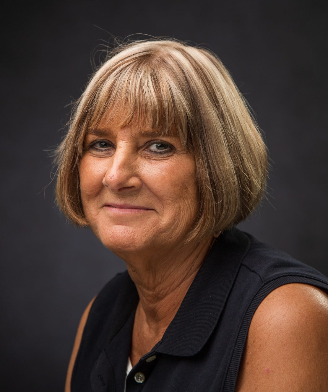 A woman with short, light brown hair and bangs, wearing a sleeveless dark collared shirt, smiles softly while looking at the camera against a dark, plain background.