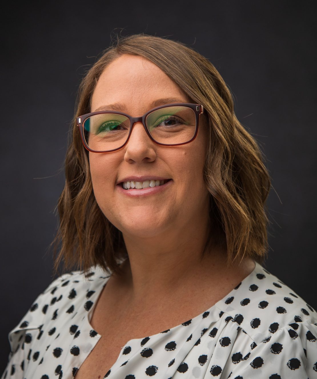 A woman with shoulder-length wavy brown hair, glasses, and a white blouse with black polka dots smiles in front of a dark background.