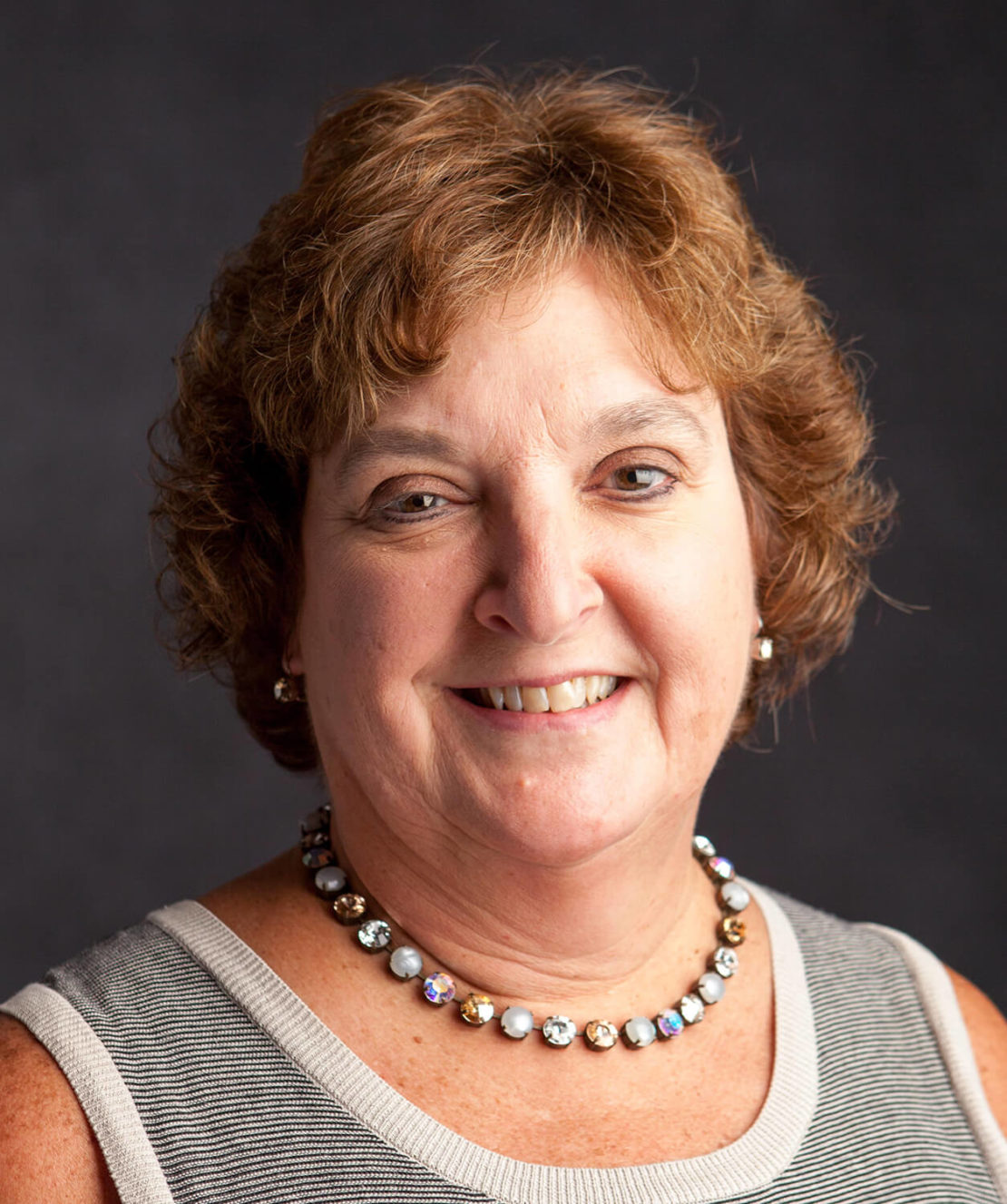 A woman with short, curly brown hair smiles at the camera. She is wearing a sleeveless gray top and a beaded necklace, with a dark, plain background behind her.
