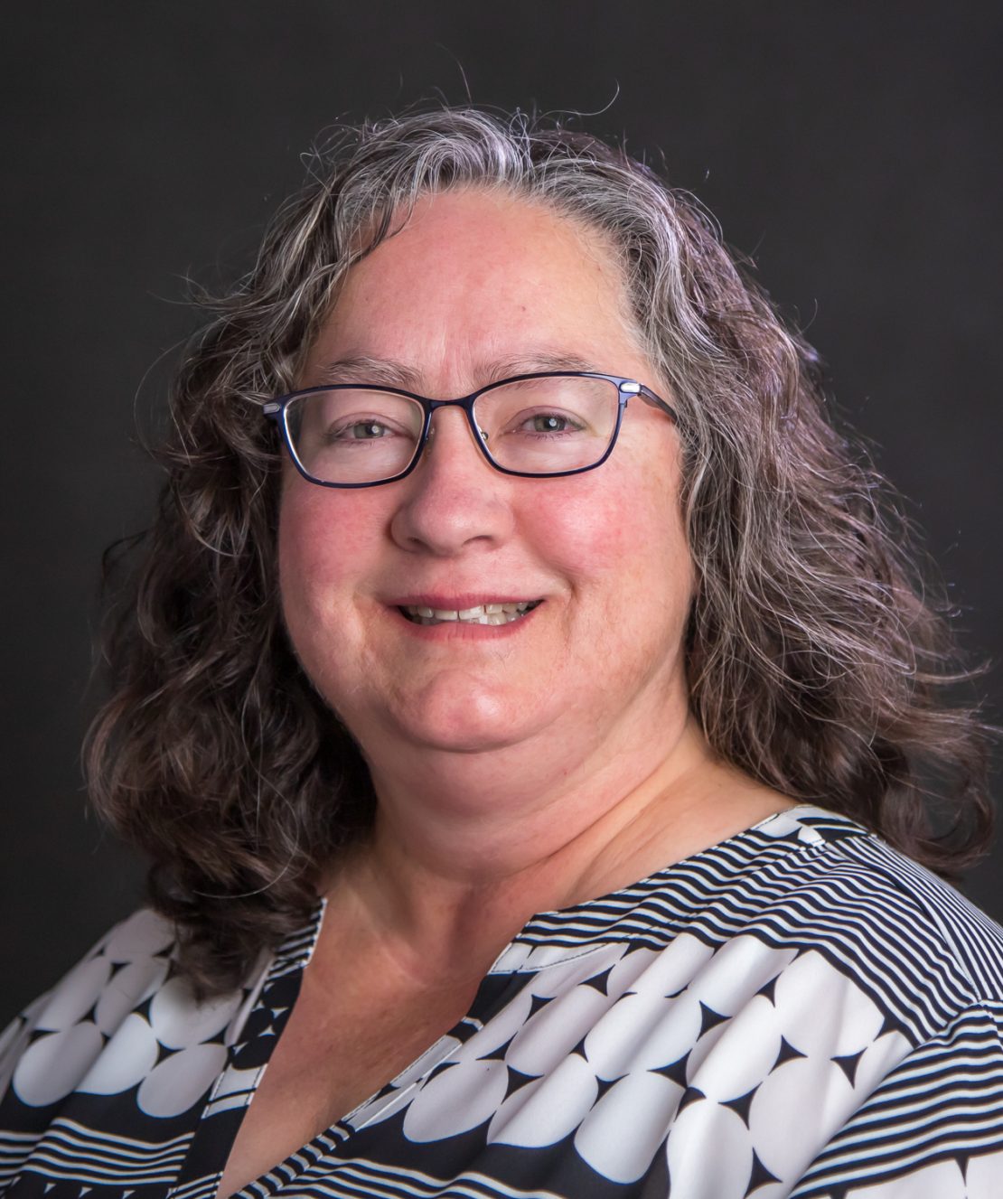 A smiling woman with wavy gray hair and glasses, wearing a patterned blouse with black and white shapes, poses in front of a dark background.