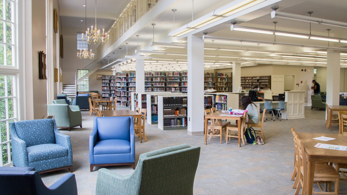 A bright library with large windows, bookshelves, wooden tables and chairs. A few people are reading or studying. Blue and green armchairs are arranged near the windows. Chandeliers hang from the ceiling.