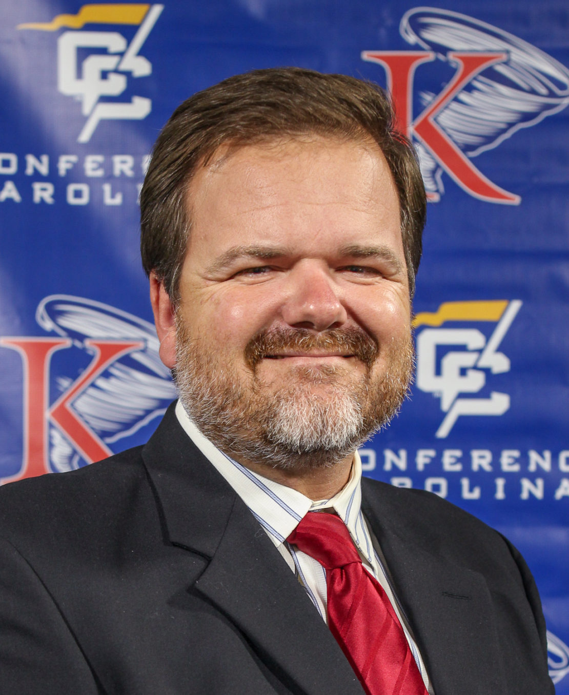 A man with short brown hair and a beard, wearing a dark suit, white shirt, and red tie, smiles in front of a blue backdrop with logos and text that read Conference Carolinas.