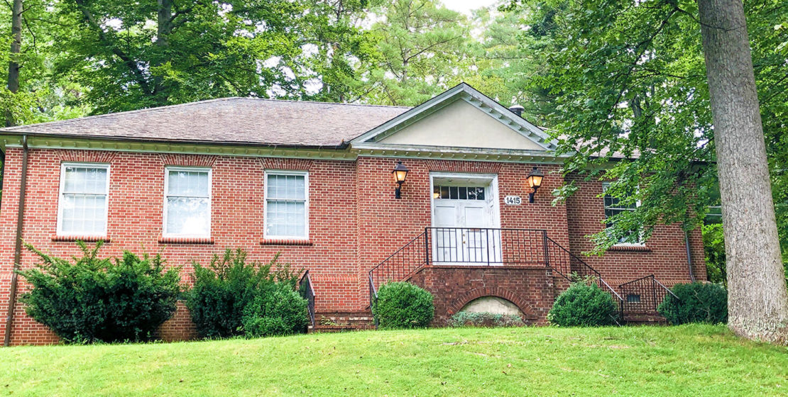 A single-story red brick building with three front windows, a white door, steps leading up to the entrance, and green bushes and trees surrounding the grassy yard.