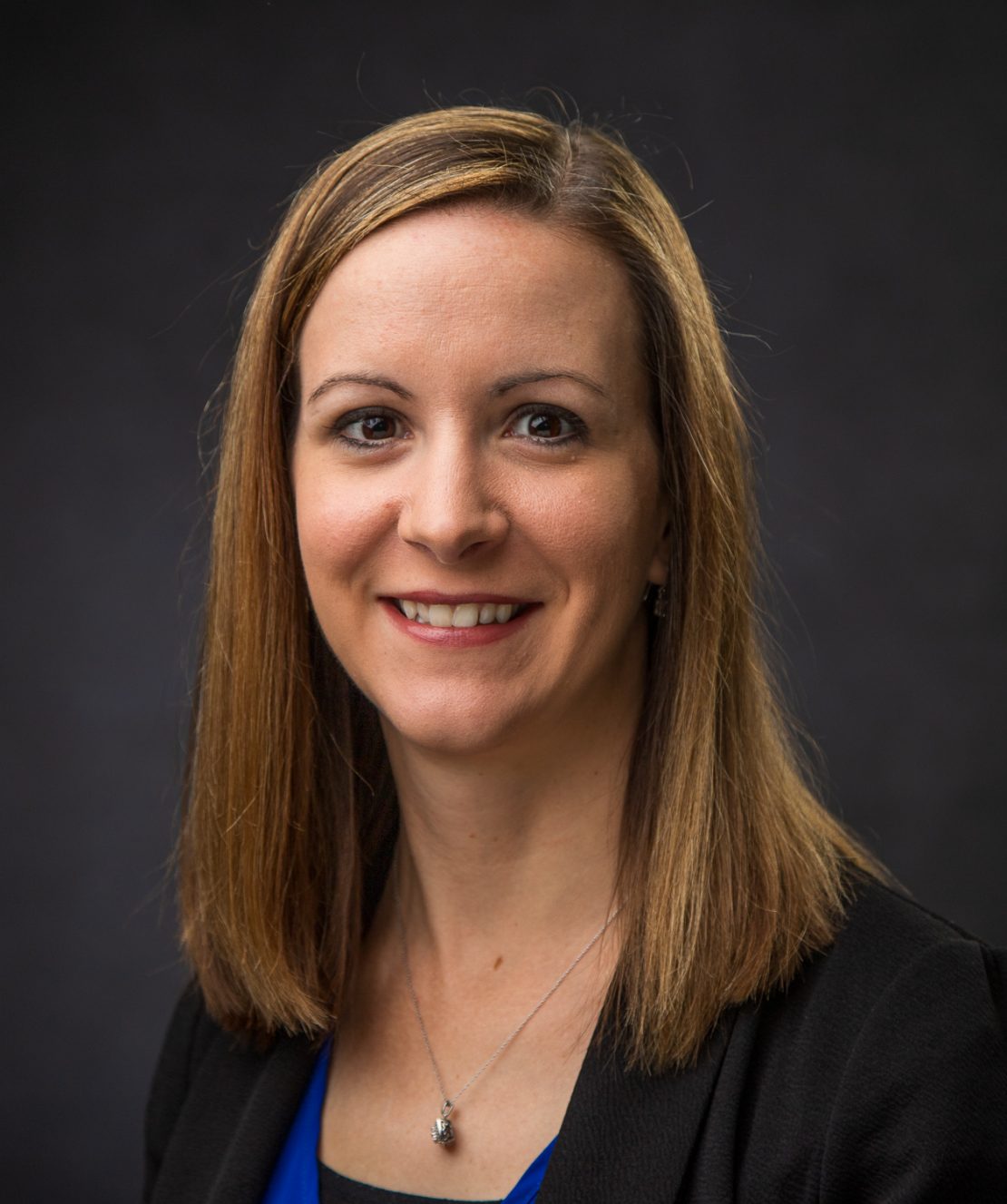 A woman with straight, shoulder-length light brown hair wearing a black blazer, blue top, and a silver necklace smiles in front of a dark, plain background.