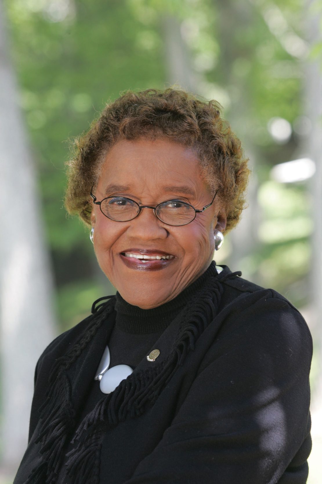 An older woman with short curly hair, glasses, and pearl earrings smiles outdoors. She is wearing a black outfit and a necklace, with green trees blurred in the background.